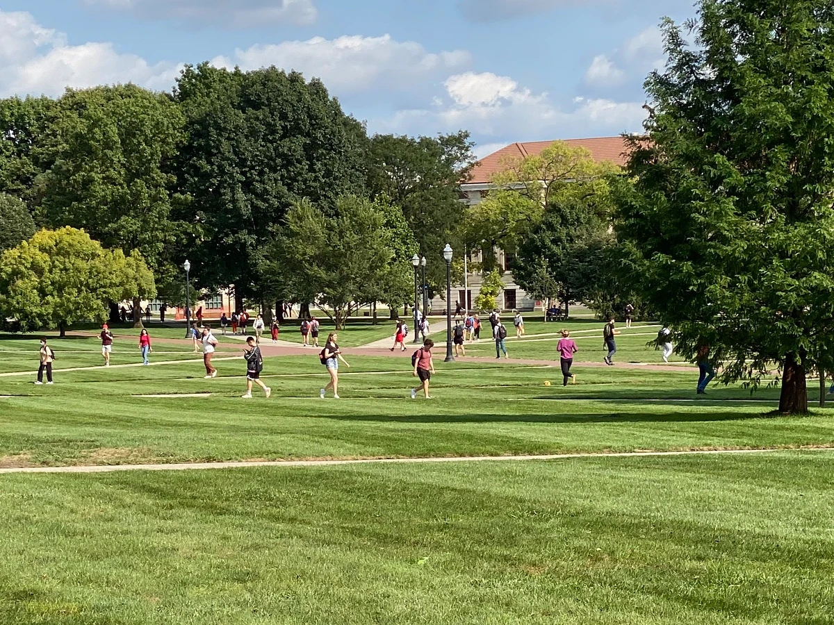 People are walking and gathering on a large, green campus under a partly cloudy sky.