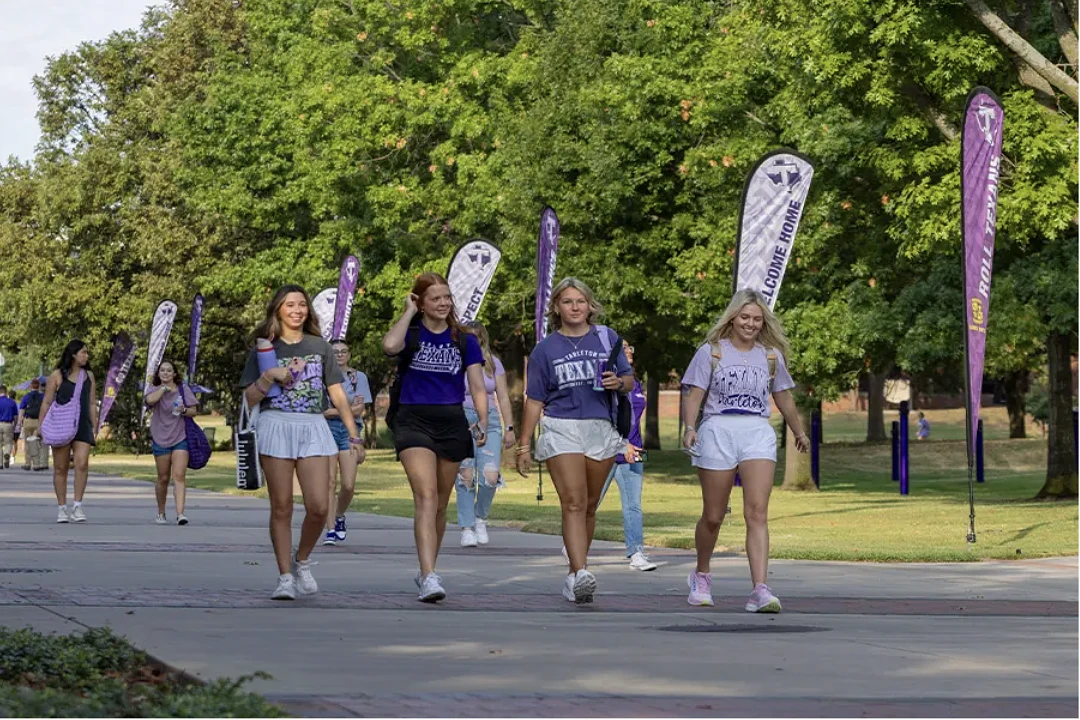 A group of young people are walking along a campus pathway lined with trees and purple flags.