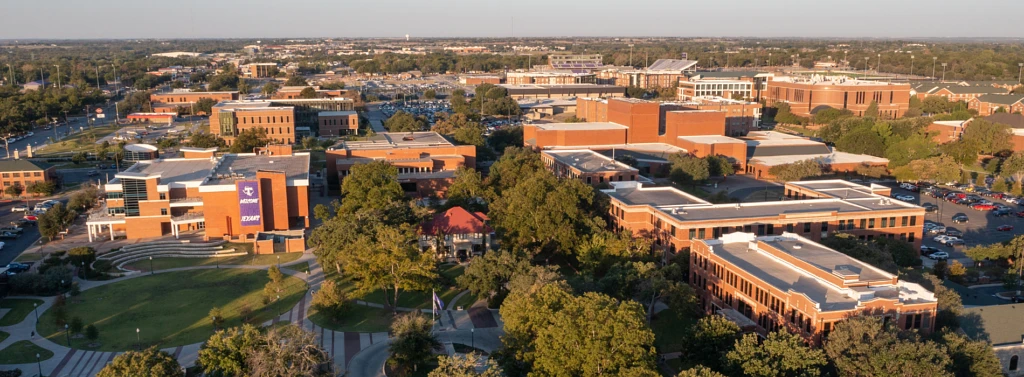 The image shows an aerial view of a large university campus with numerous brick buildings surrounded by green trees and pathways.