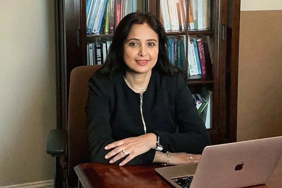 A person sits at a desk with a laptop, smiling, in front of a bookshelf filled with books.