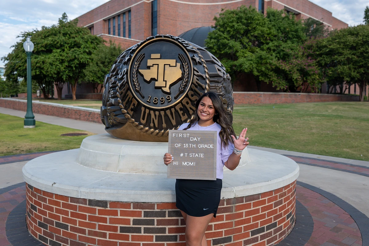 A smiling person stands in front of a university ring sculpture holding a sign that reads, "First day of 13th grade #TState Hi Mom!"