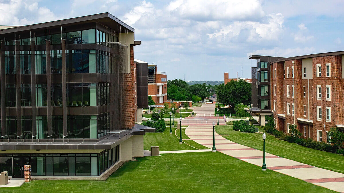 A modern campus with brick and glass buildings flanking a tree-lined walkway under a partly cloudy sky.