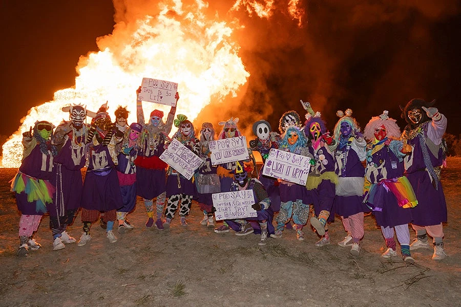 A group of people dressed in colorful costumes and masks pose with signs in front of a large bonfire.
