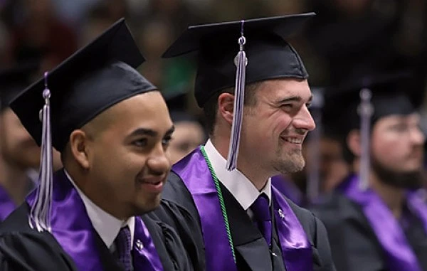 Two graduates wearing caps and gowns with purple sashes sit smiling during a ceremony.